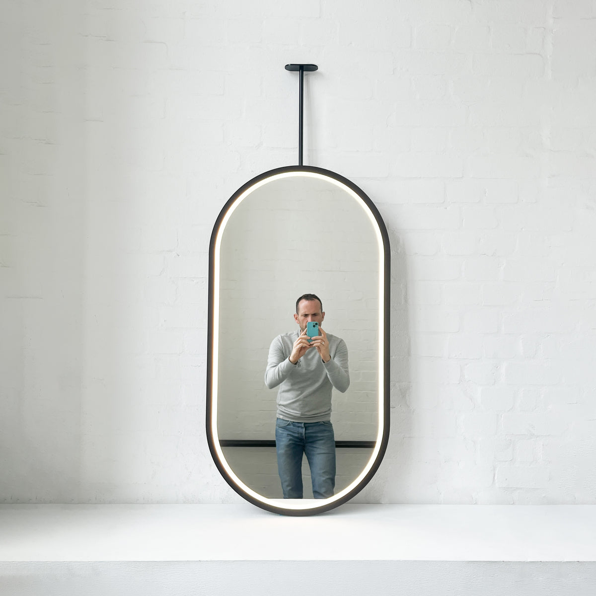 Modern front illuminated Capsula™ ceiling suspended pill shaped mirror with a black powder coated full frame.