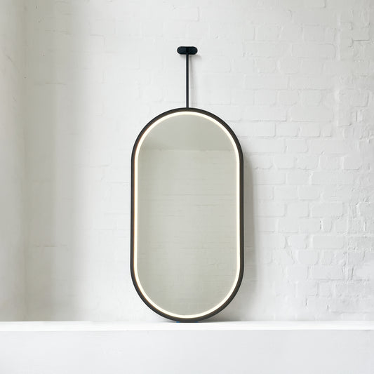 Modern front illuminated Capsula™ ceiling suspended pill shaped mirror with a black powder coated full frame