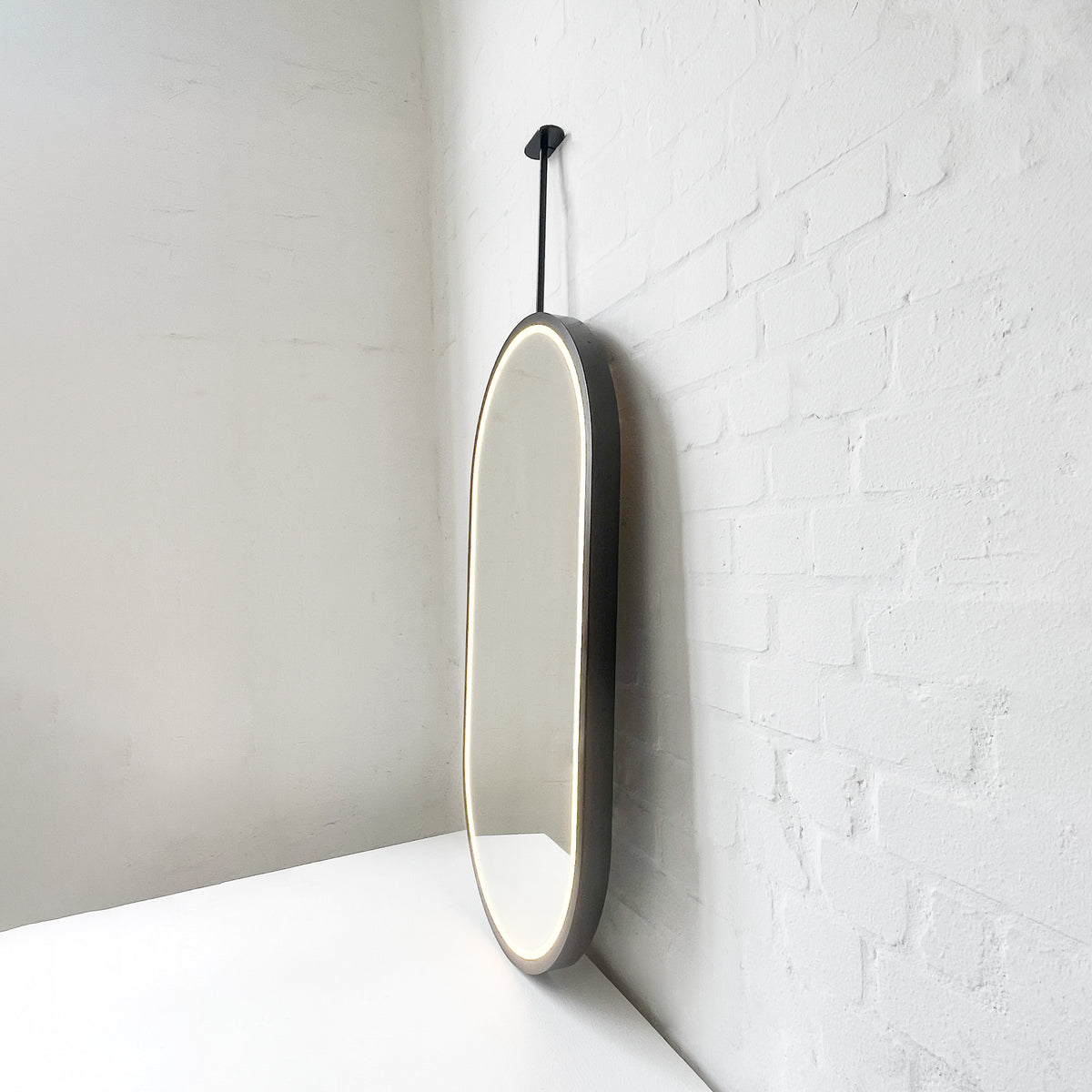 Modern front illuminated Capsula™ ceiling suspended pill shaped mirror with a black powder coated full frame.