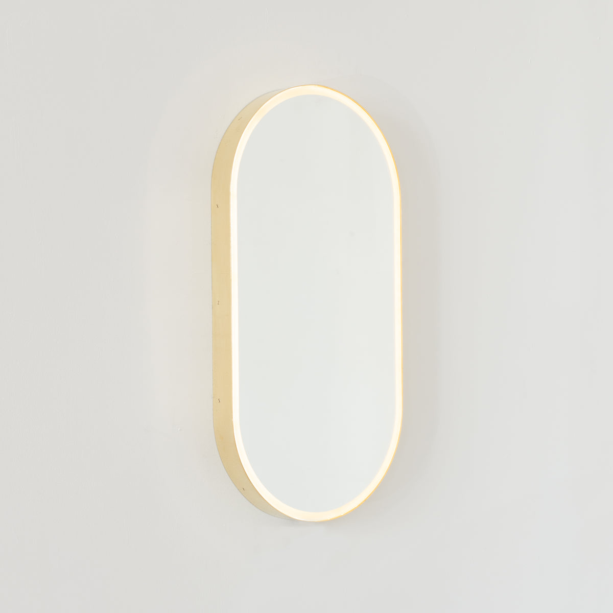 Modern pill shaped mirror with a minimalist brushed brass frame and front illumination