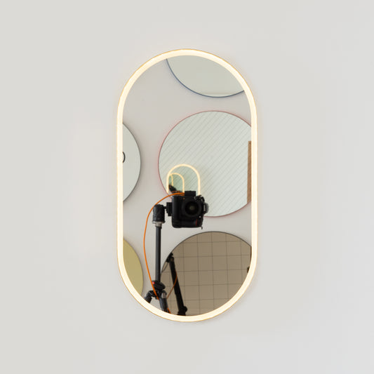 Modern pill shaped mirror with a minimalist brushed brass frame and front illumination